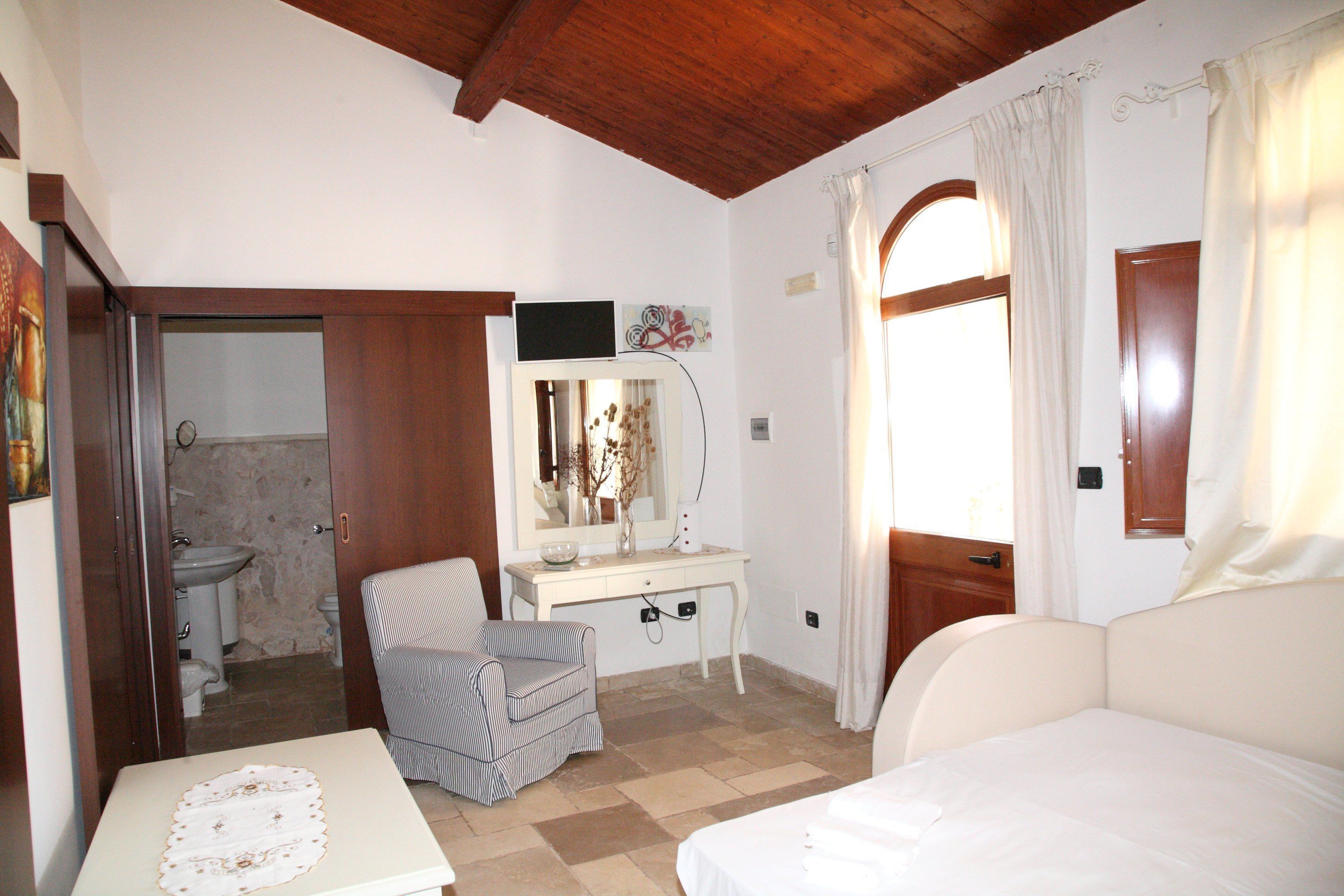 Une chambre à coucher comprenant un lit, une table de chevet, une chaise, un bureau avec un miroir, et une salle de bain visible à travers une porte ouverte.