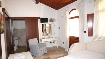 Une chambre à coucher comprenant un lit, une table de chevet, une chaise, un bureau avec un miroir, et une salle de bain visible à travers une porte ouverte.