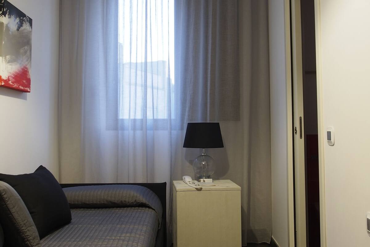 A hotel room with a bed, a bedside table with a lamp, a window with curtains, and a door.