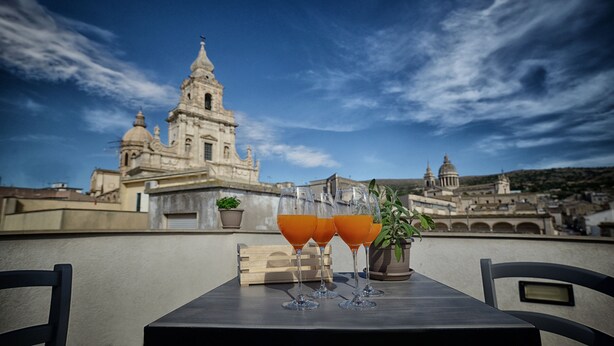 A table with three glasses of orange juice, a wooden block, and a potted plant on a rooftop terrace with a view of a historic church and cityscape. - Terrazzani Suite (Comiso)