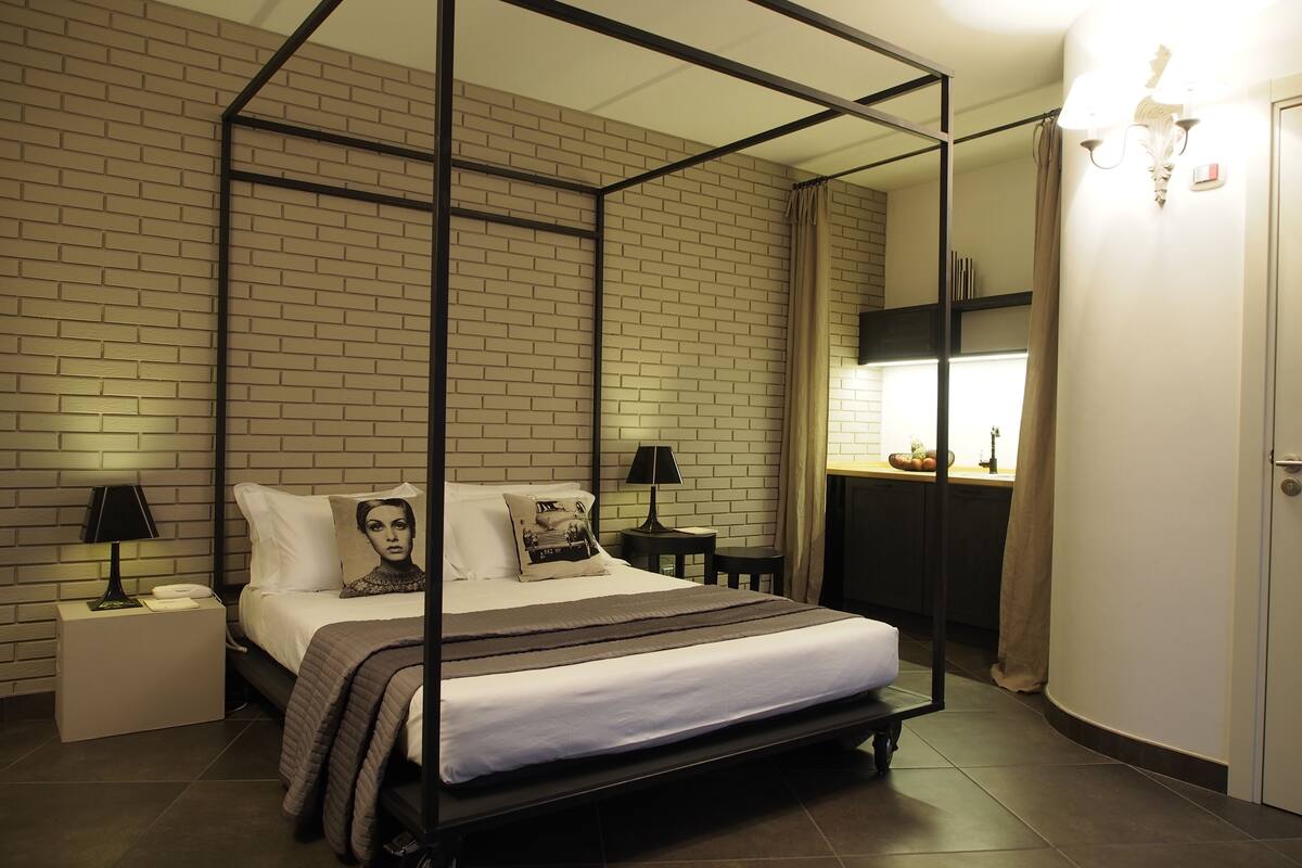 A four-poster bed with a white mattress and a brown blanket, a nightstand with a lamp, a small table with a lamp, a mirror, and a wardrobe.