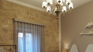 A bedroom with a large bed, a bedside table, a nightstand, a hanging light fixture, a window with curtains, and a stone wall. - Terrazzani Suite (Comiso)