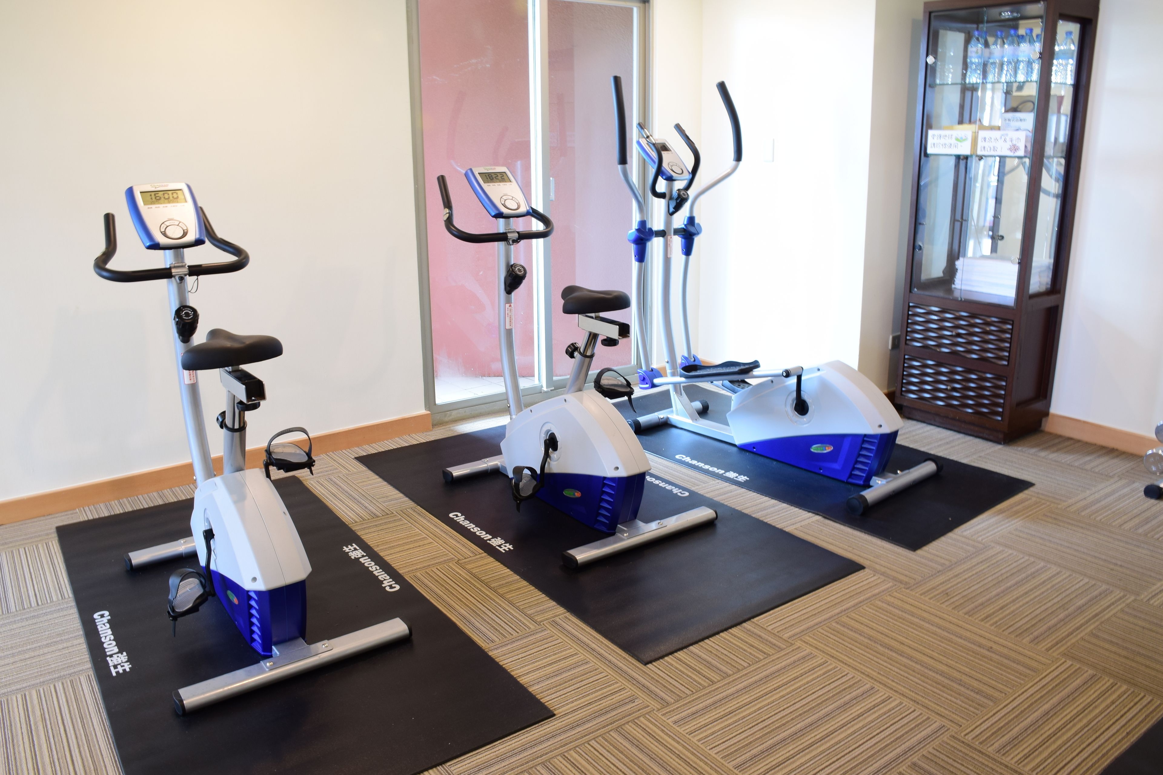 Three stationary bikes on black mats in a room with a wooden floor.