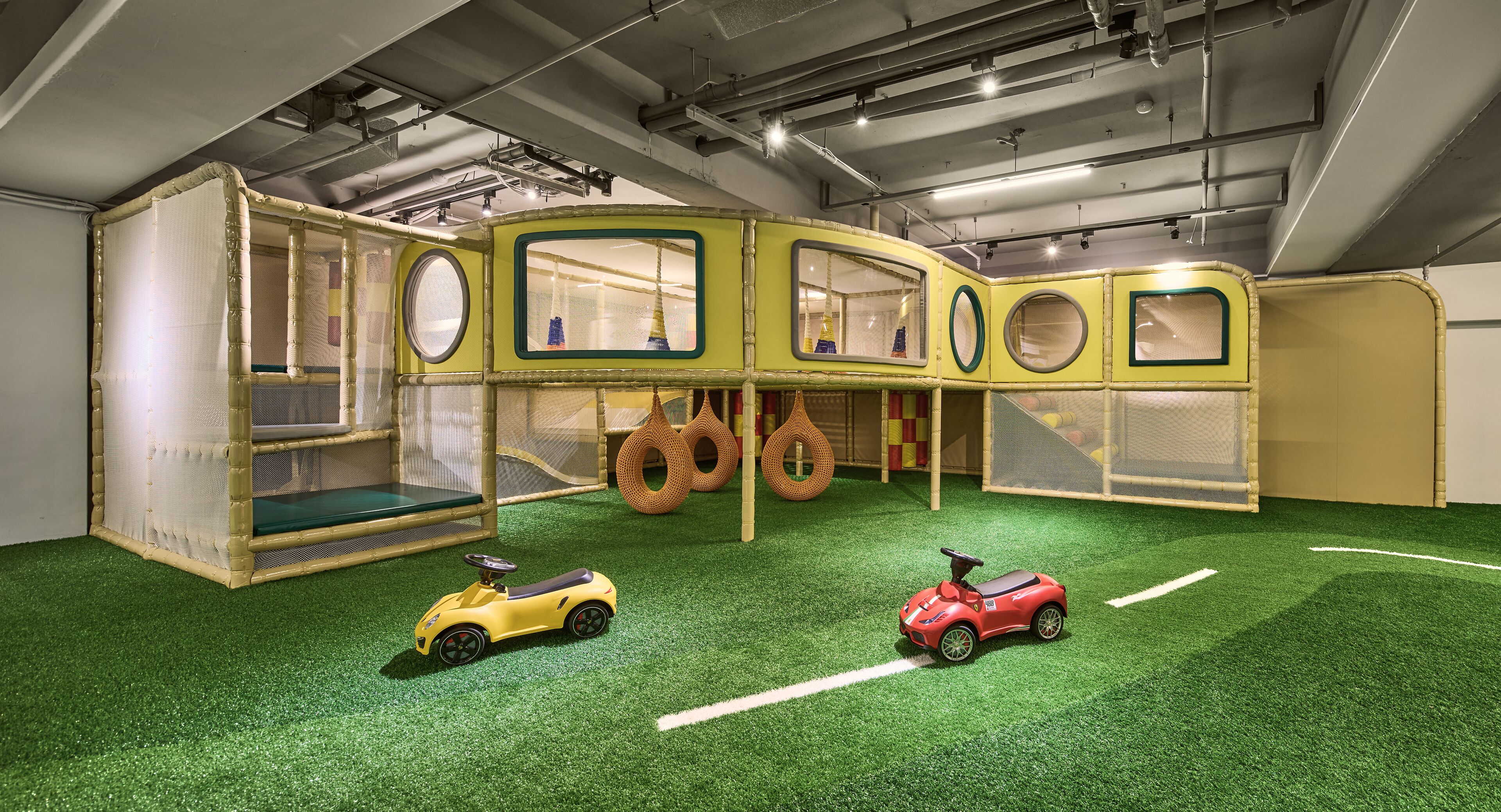A play area with a green artificial turf floor, two toy cars, and a play structure with circular windows and a netted enclosure.