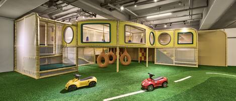 A play area with a green artificial turf floor, two toy cars, and a play structure with circular windows and a netted enclosure.
