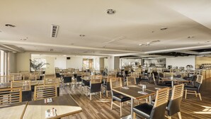 A spacious restaurant with wooden tables and chairs, a kitchen area in the background, and large windows allowing natural light.