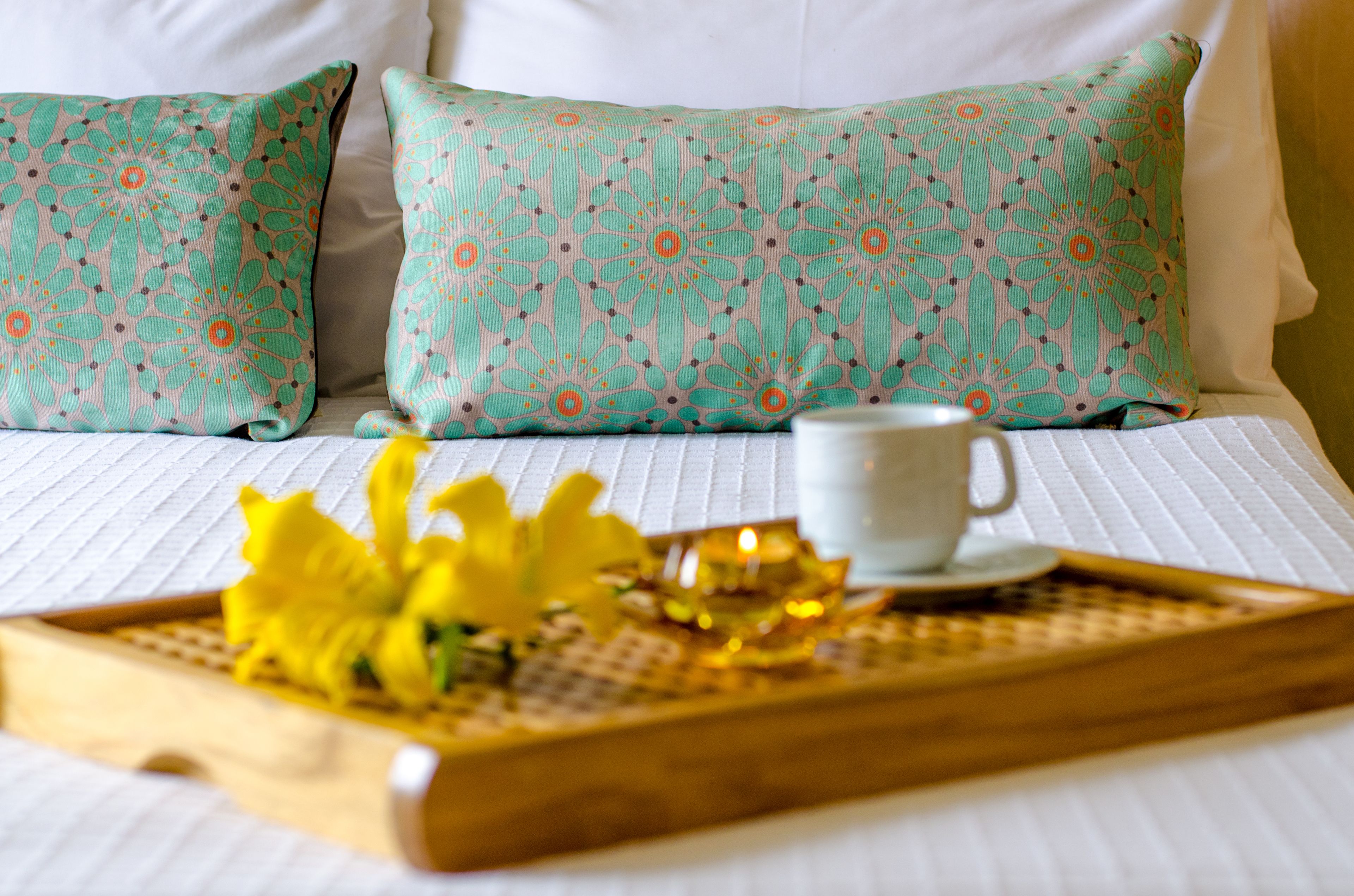 Una cama bien tendida con ropa blanca, dos almohadas estampadas en color aguamarina y una bandeja con una taza blanca y flores amarillas.