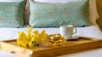 Una cama bien tendida con ropa blanca, dos almohadas estampadas en color aguamarina y una bandeja con una taza blanca y flores amarillas.