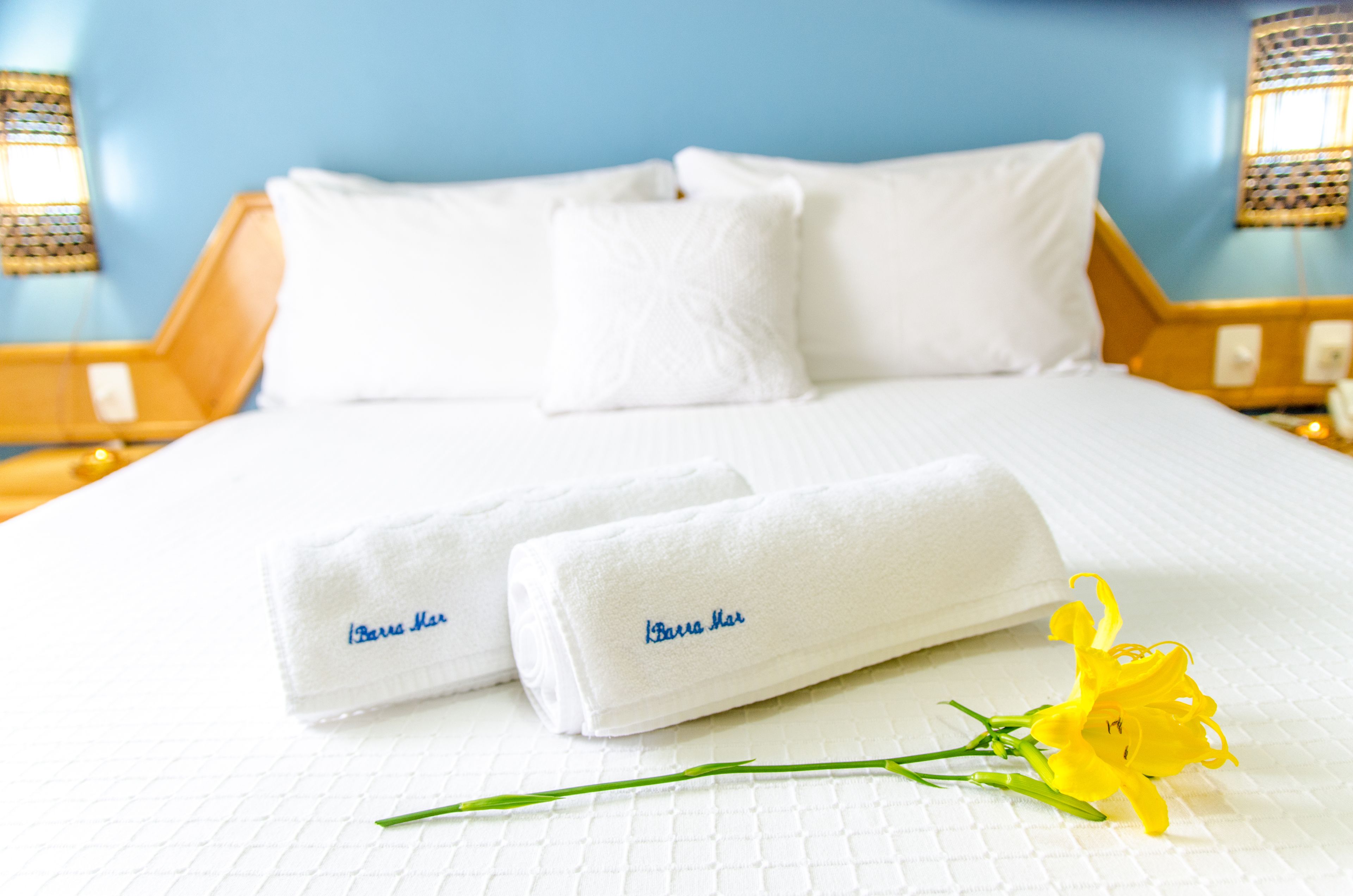 A neatly made bed with white linens and two towels with "Barra Mar" embroidered on them, a yellow flower on the bed, and a blue wall in the background.