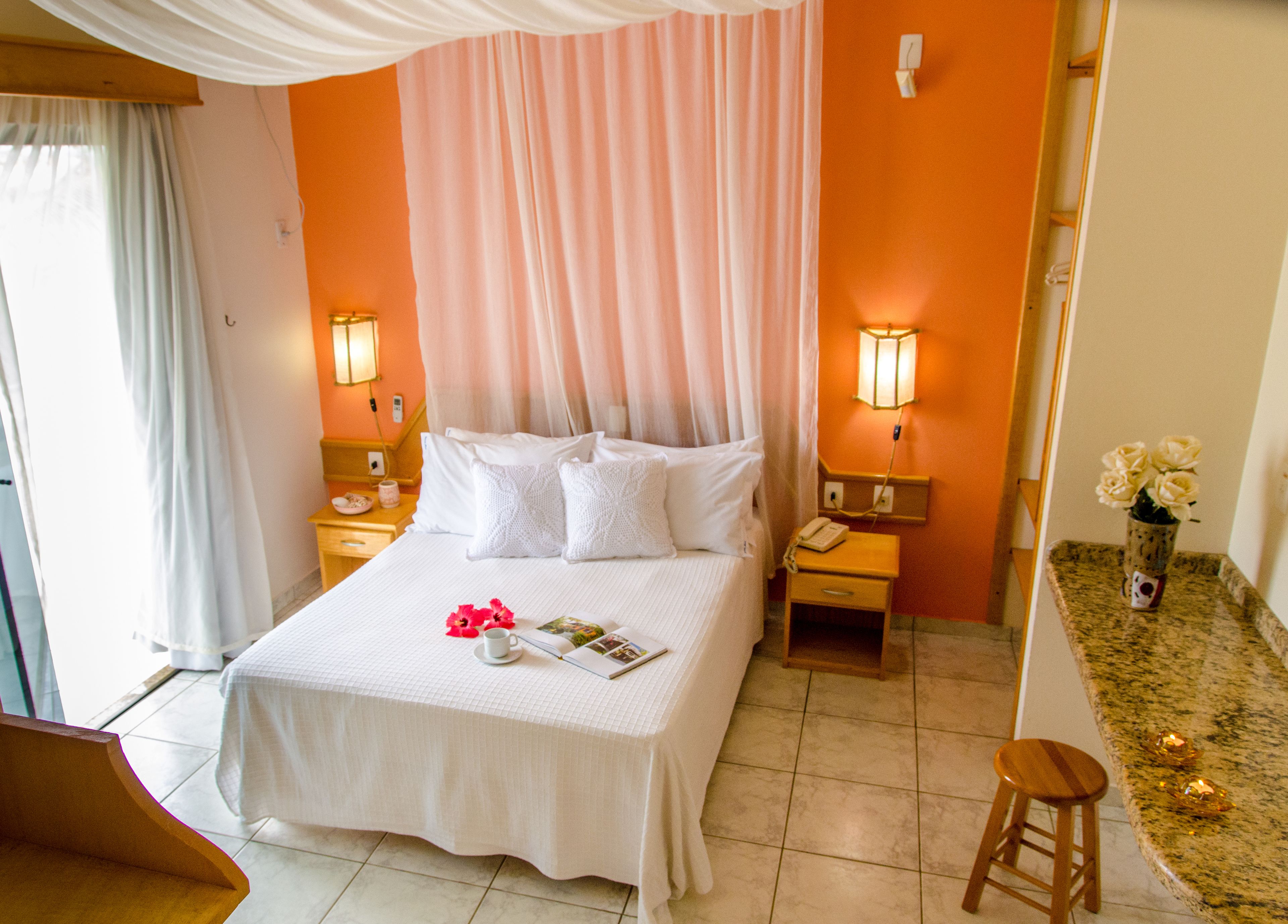 Una habitació d'hotel amb un llit gran, dues taules de nit, un taulell, un tamboret de fusta i una taula petita amb un vas de flors.