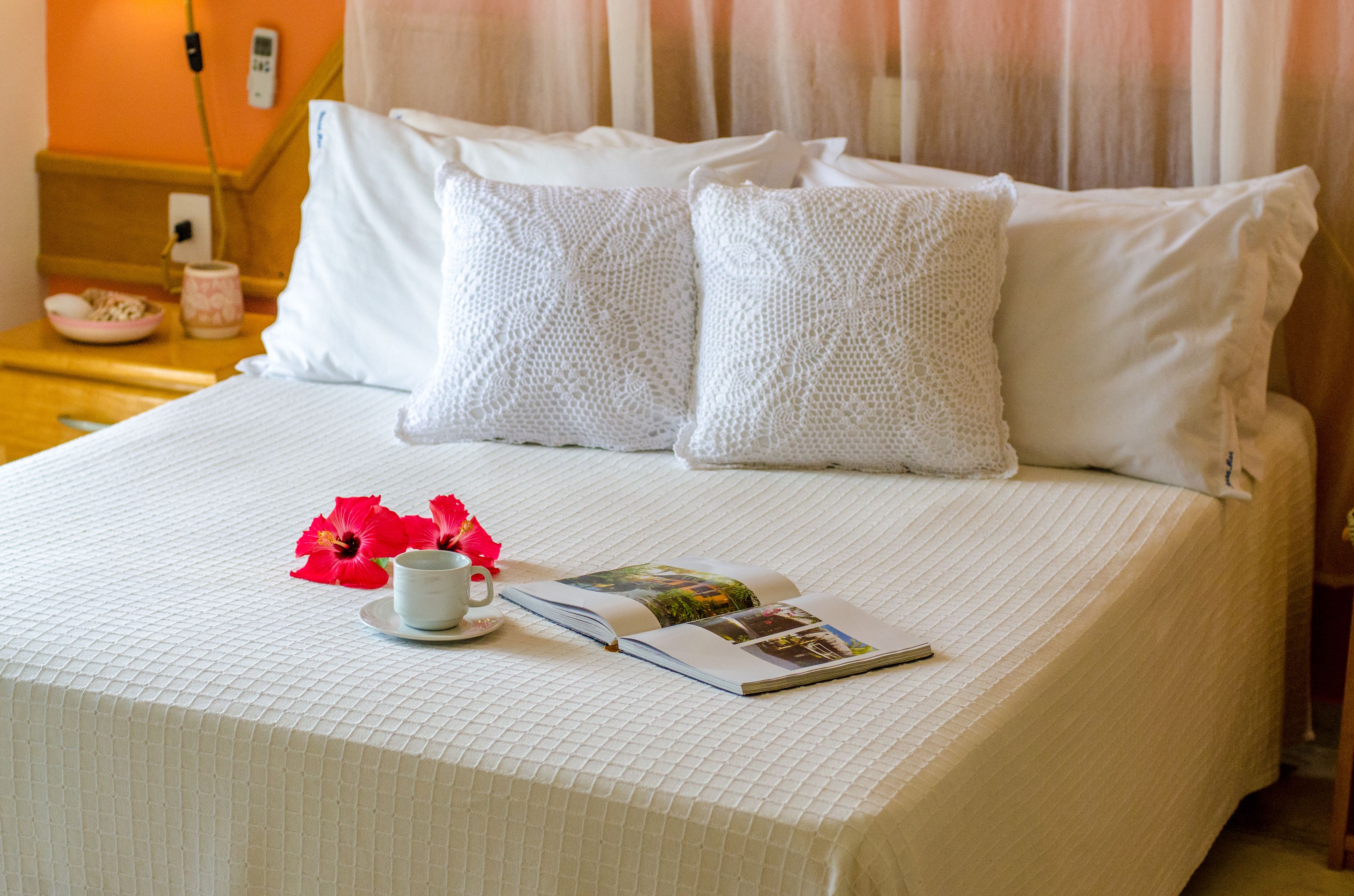 Una cama bien hecha con sábanas blancas y almohadas decorativas, una mesita con una taza y un libro, y un frutero a un lado.
