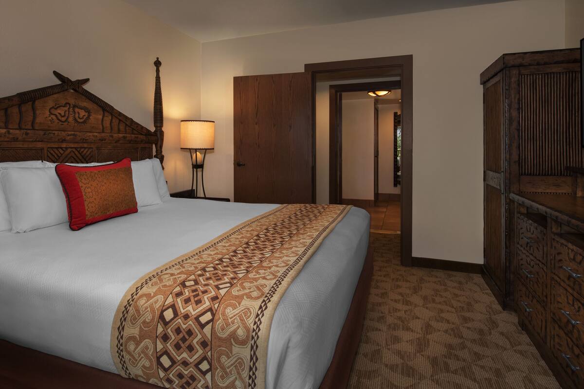 A hotel room with a large bed, a wooden headboard, a red accent pillow, a patterned throw blanket, a nightstand with a lamp, a wooden wardrobe, and a television.