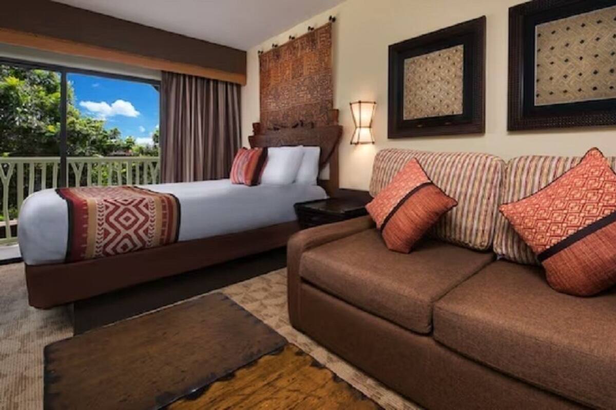 A hotel room with a bed, a sofa, a nightstand, a lamp, and a view of the outdoors.