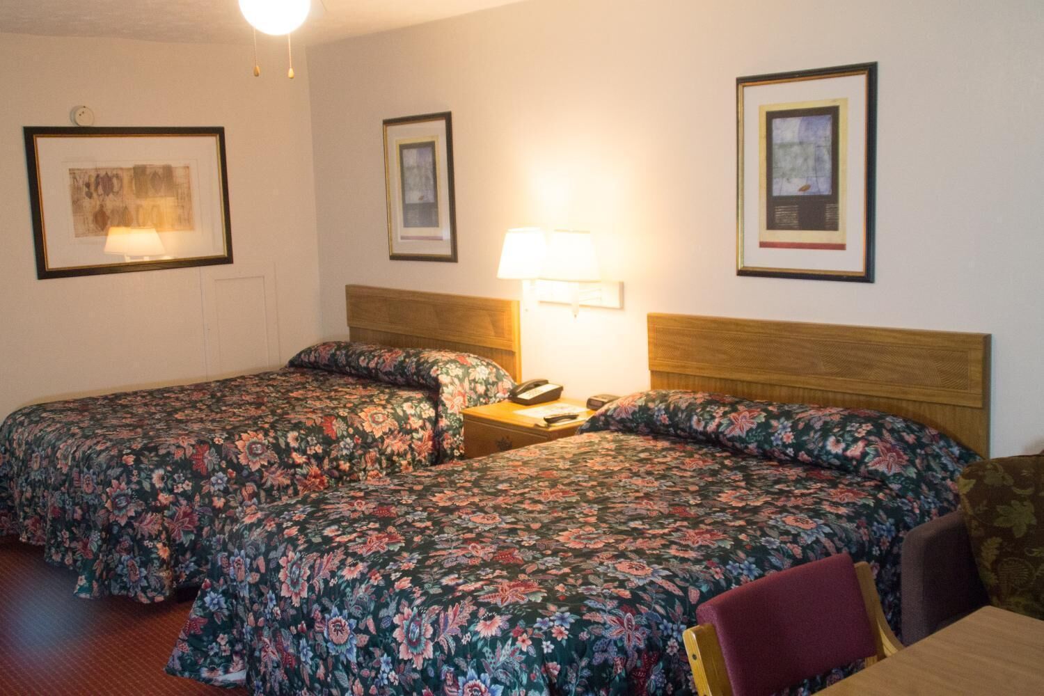 A hotel room with two single beds, a wooden headboard, a bedside table with a lamp, and framed pictures on the walls.