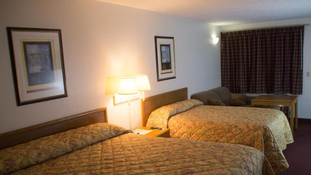 A hotel room with two single beds, a nightstand with a lamp, a small table, and a window with curtains. - Tazewell Motor Lodge (Tazewell)