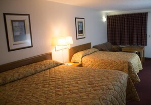 A hotel room with two single beds, a nightstand with a lamp, a small table, and a window with curtains. - Tazewell Motor Lodge (Tazewell)