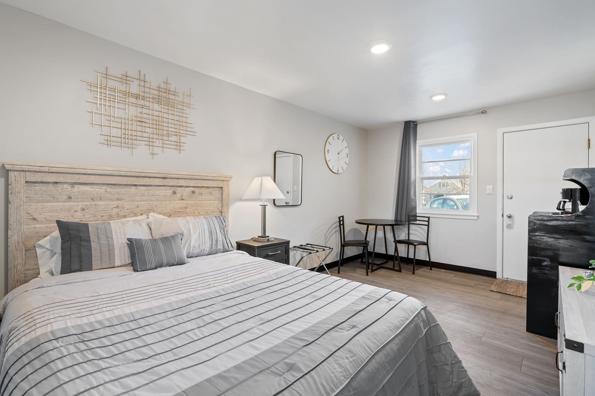 A hotel room with a large bed, a small desk, a chair, a clock, and a window with a view.