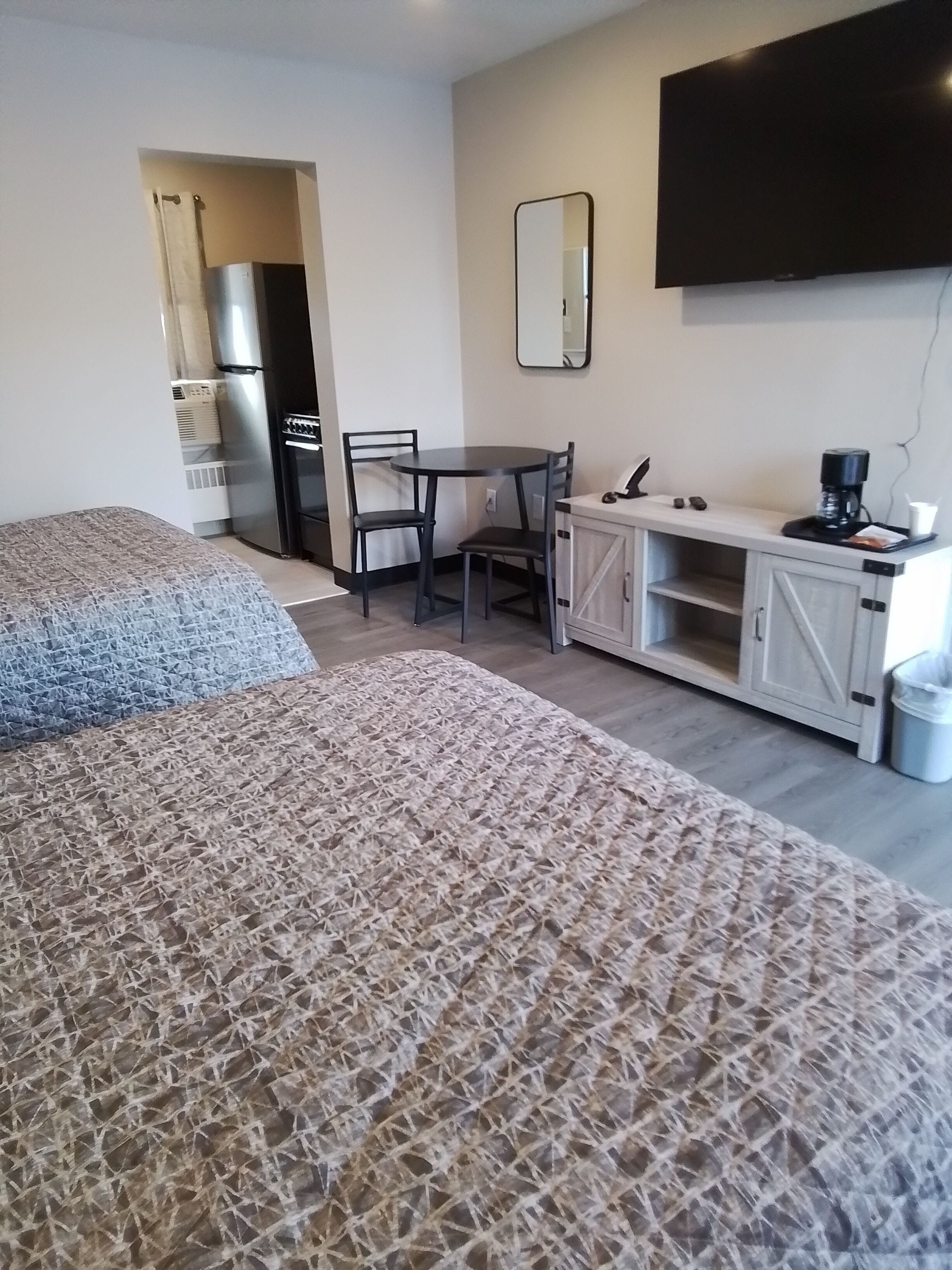 A bedroom with two beds, a small kitchenette, a dining table with chairs, a TV, and a trash bin.