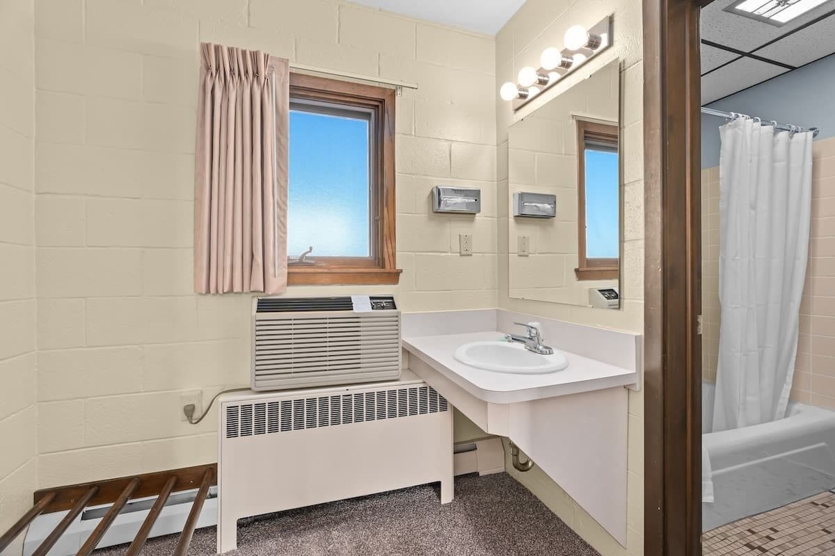 A compact bathroom with a white sink, a mirror, a window with curtains, a heater, and a shower area.