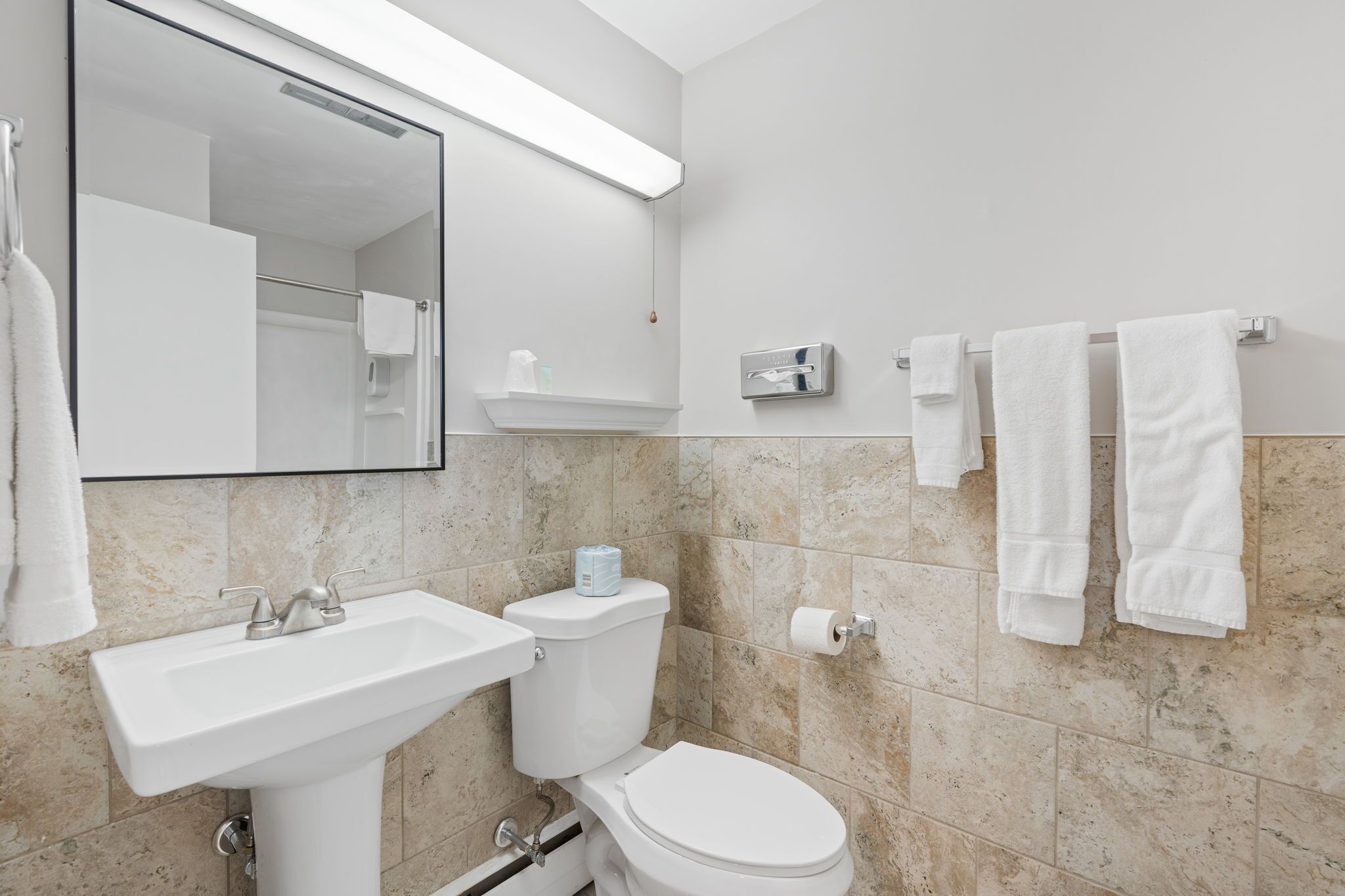 A bathroom with a white sink, toilet, and a mirror. There are towels hanging on the wall and a shelf with toiletries.
