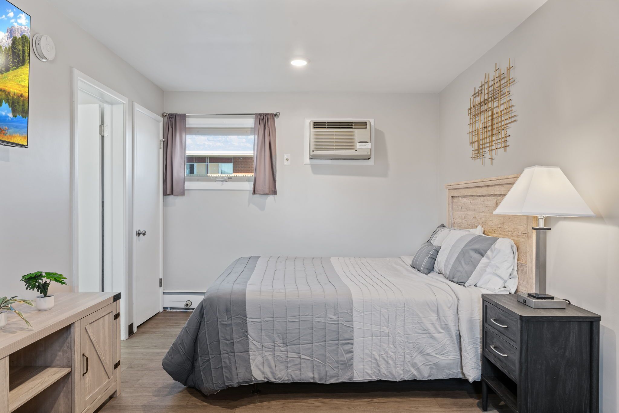 A bedroom with a bed, bedside table, lamp, air conditioning unit, and a window with curtains.