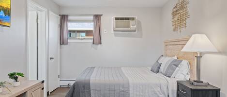Un dormitorio con cama, mesita de noche, lámpara, equipo de aire acondicionado y ventana con cortinas.