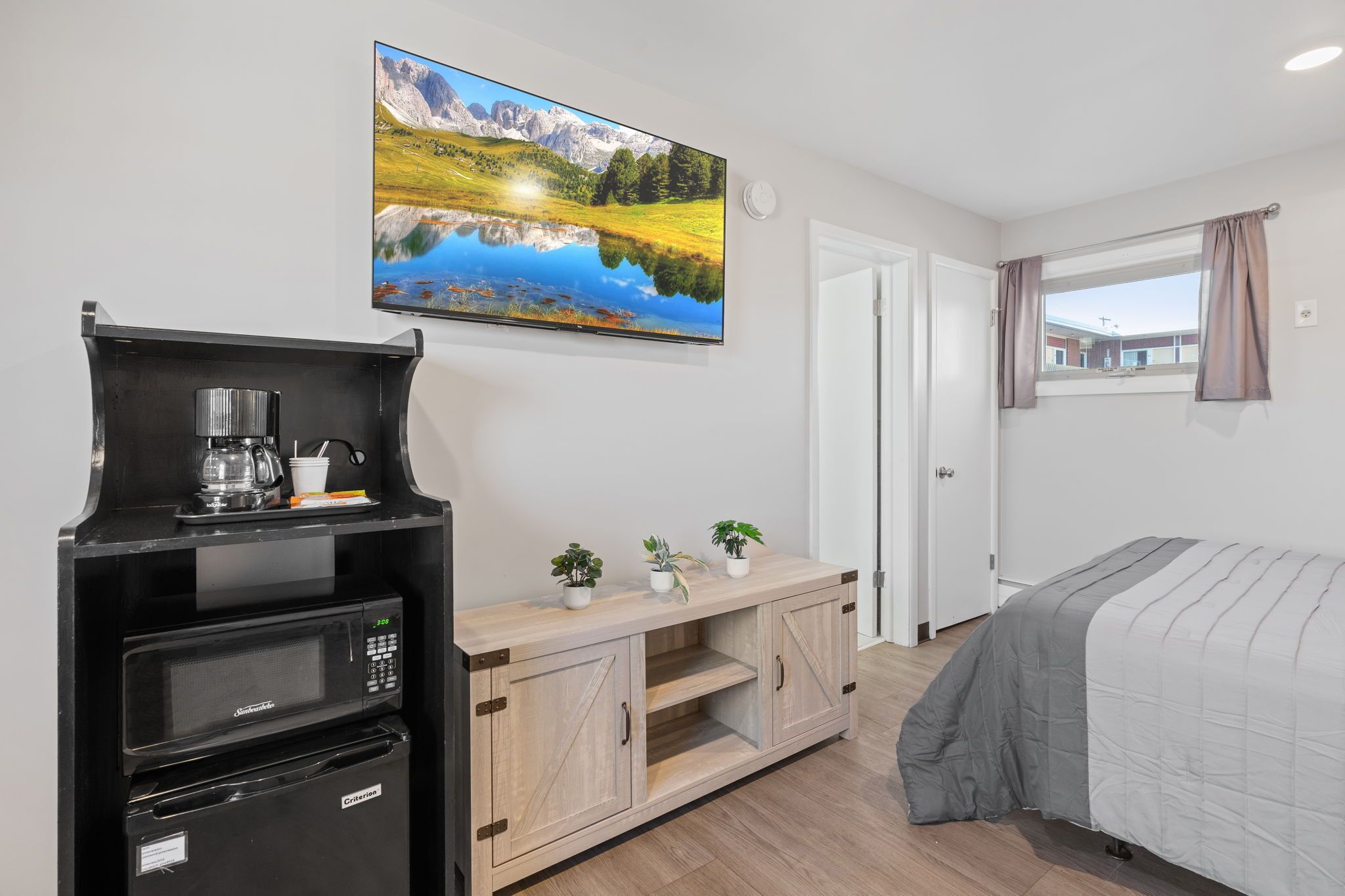 Een slaapkamer met een bed, een tv aan de muur, een kitchenette met een koffiezetapparaat, en een raam met gordijnen.