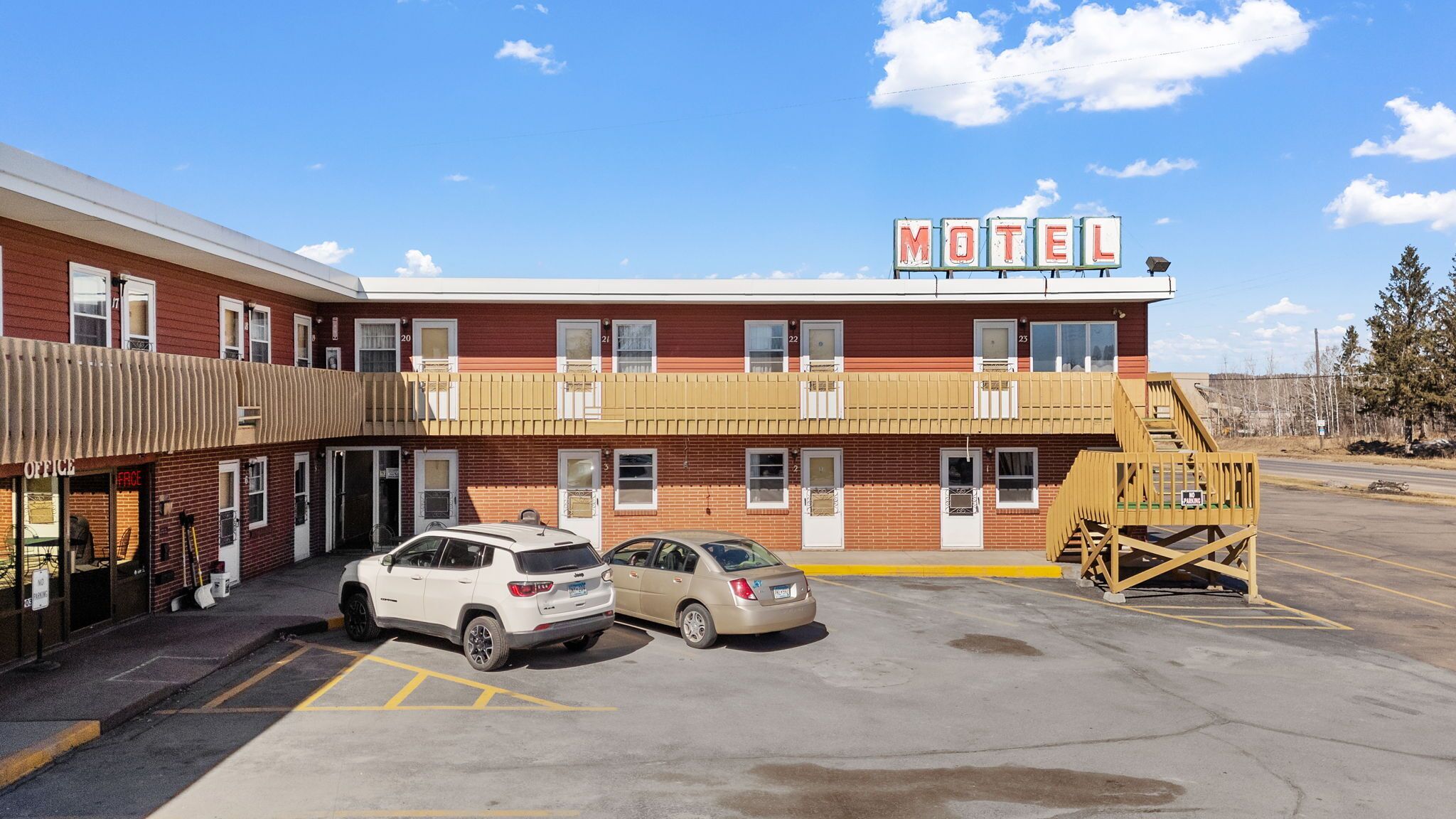 A motel with a two-story building, a parking lot, and a small outdoor area with a wooden structure.