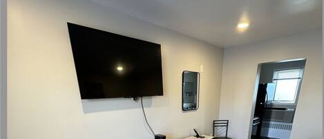 A compact room with a flat-screen TV mounted on the wall, a small round table with chairs, a kitchenette area, and a bed with a patterned cover.