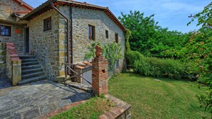 A stone building with a brick chimney, a set of stone steps leading to a door, and a small garden area with grass and shrubs. - Montescalari (Figline e Incisa Valdarno)