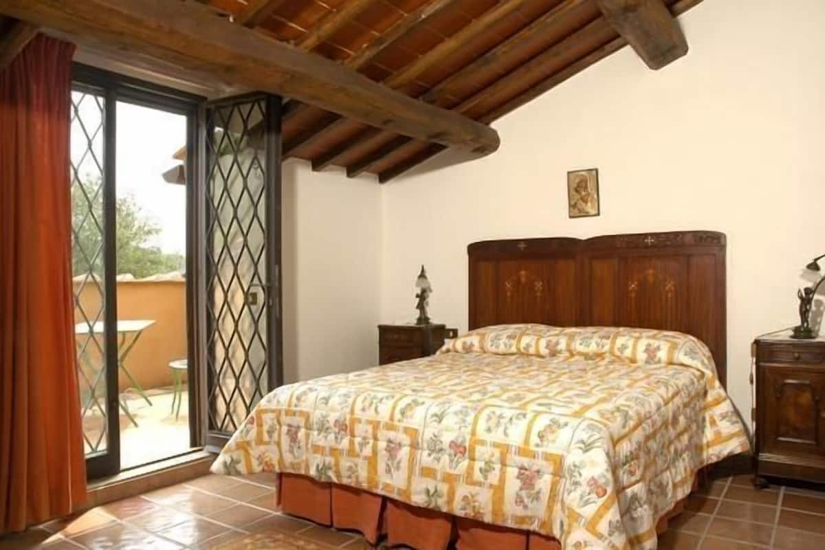 A bedroom with a wooden bed, a nightstand, a window with a view, and a door leading to an outdoor area.