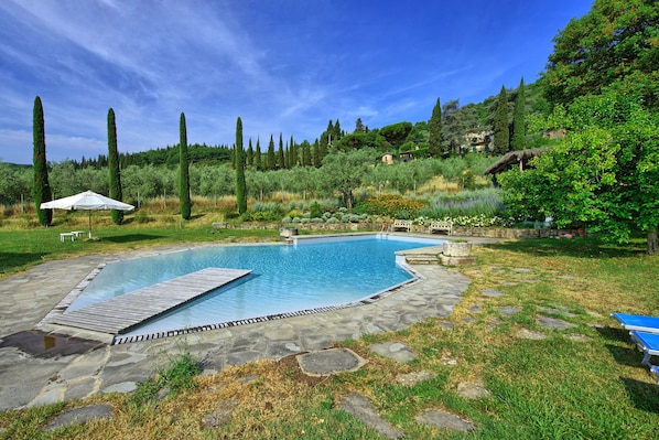 Piscina com deck de madeira, rodeada por um pátio de pedra e um jardim exuberante com diversas plantas e árvores.