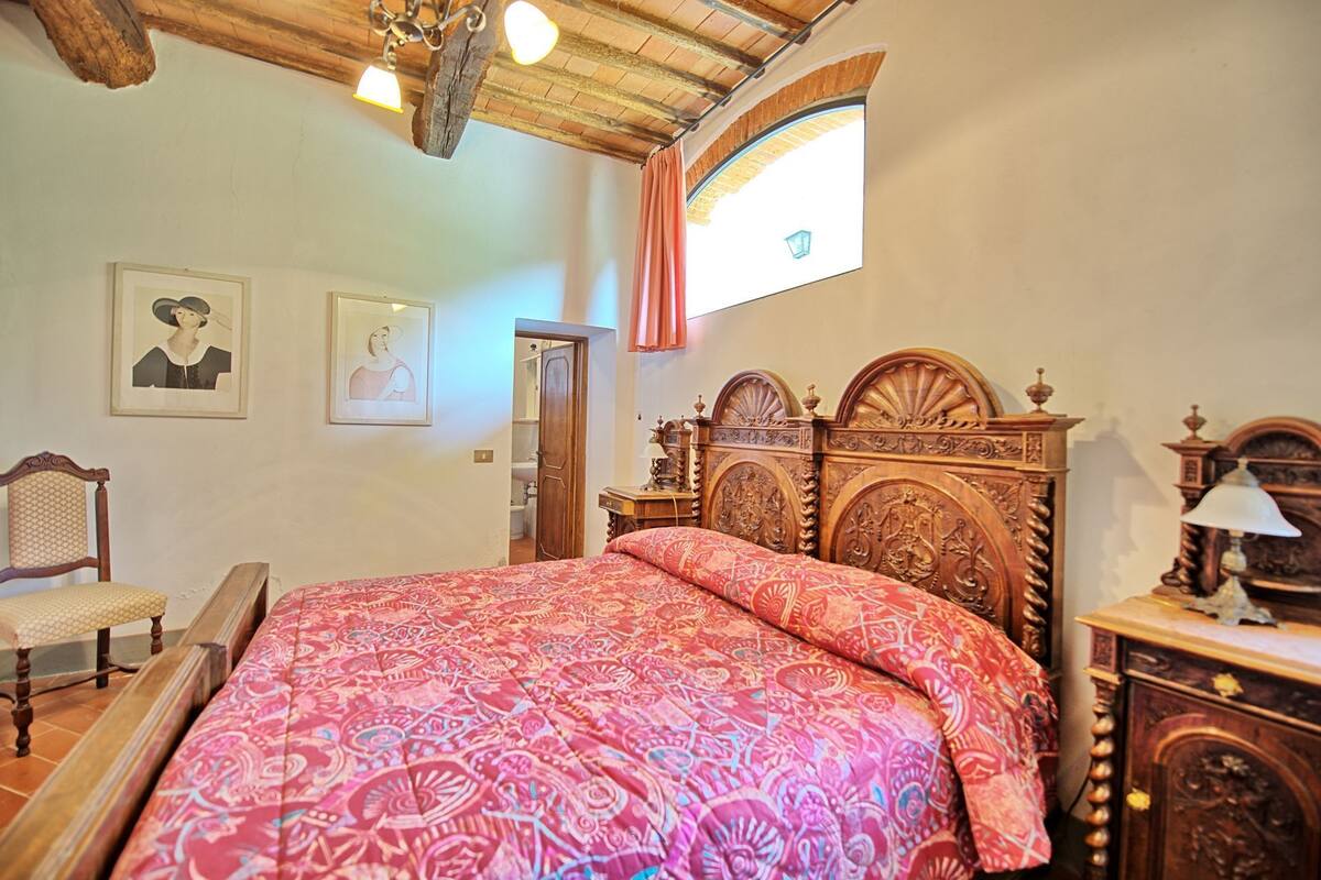 A bedroom with a large bed, wooden headboard, a chair, a nightstand, and a window with a view.