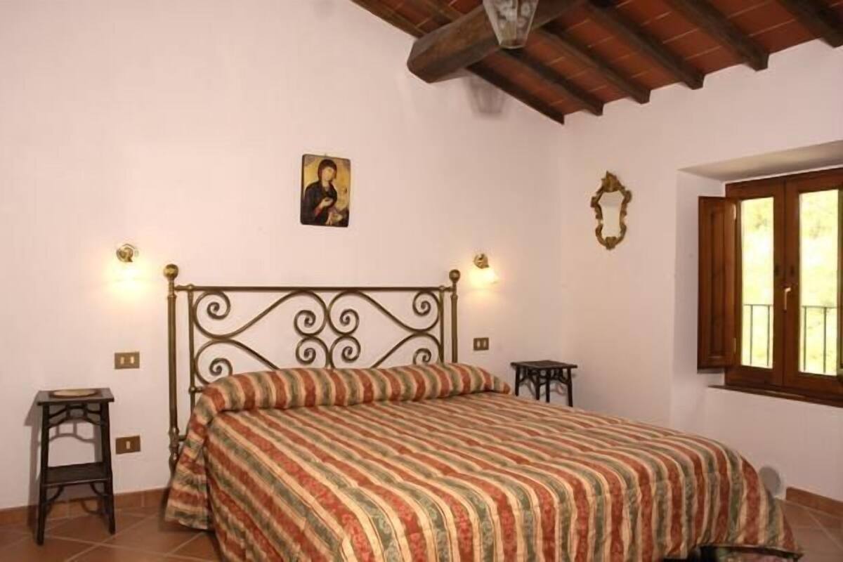 A bedroom with a metal bed frame, a striped bedspread, a small bedside table, a wall-mounted light, and a window with wooden shutters.