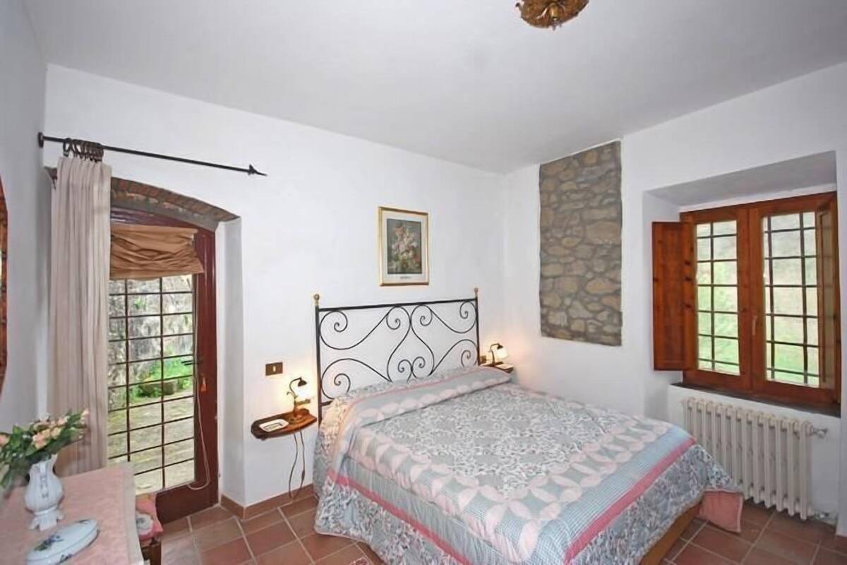 A bedroom with a bed, a nightstand, a window with curtains, a stone wall, and a radiator.