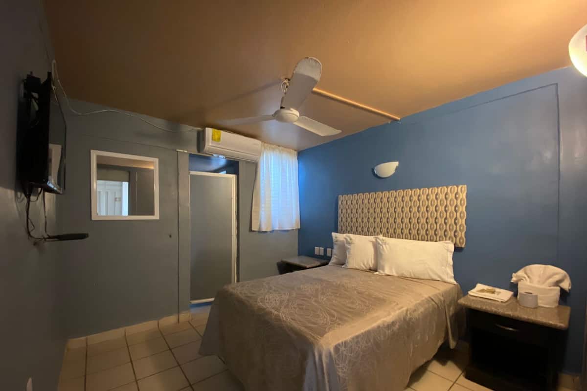 Un dormitorio con cama, ventilador de techo, mesita de noche, televisor montado en la pared y una puerta.