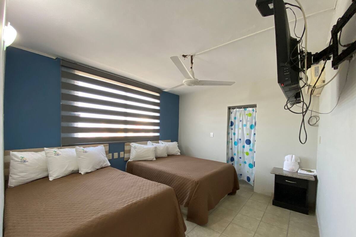 Habitación de hotel con dos camas, un ventilador de techo, una ventana con persianas, una cortina y una mesita con una taza.