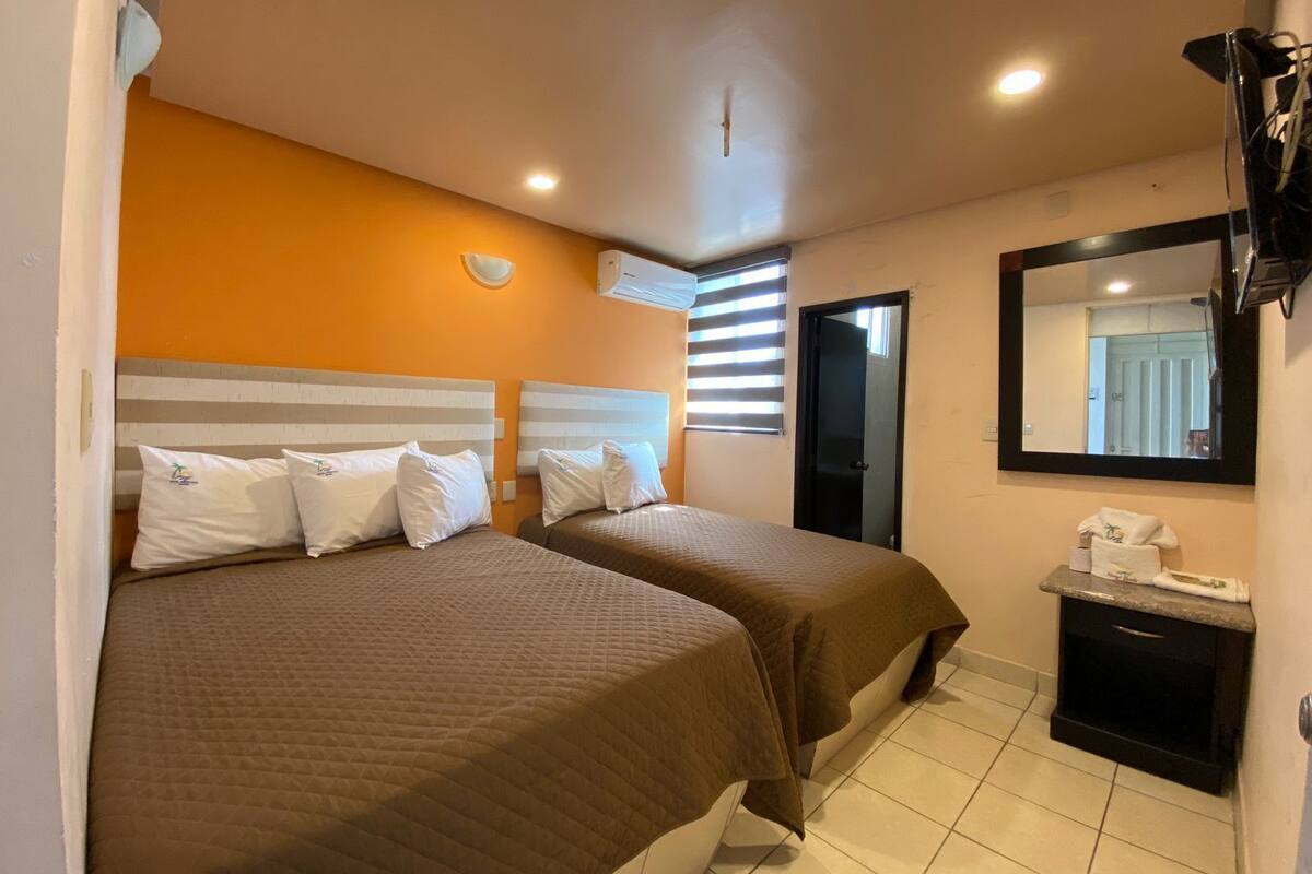 Habitación de hotel con dos camas, una mesita de noche pequeña, un espejo y una ventana con persianas.