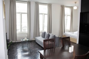 Quarto de hotel moderno com sofá cinza, mesa de jantar de madeira e pequena mesinha de apoio.