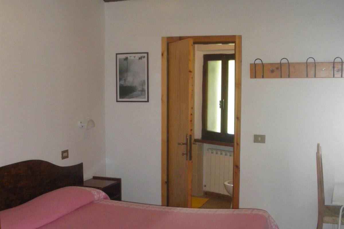 A bedroom with a bed, a wooden door, a small table, a chair, a radiator, and a wall-mounted coat rack.