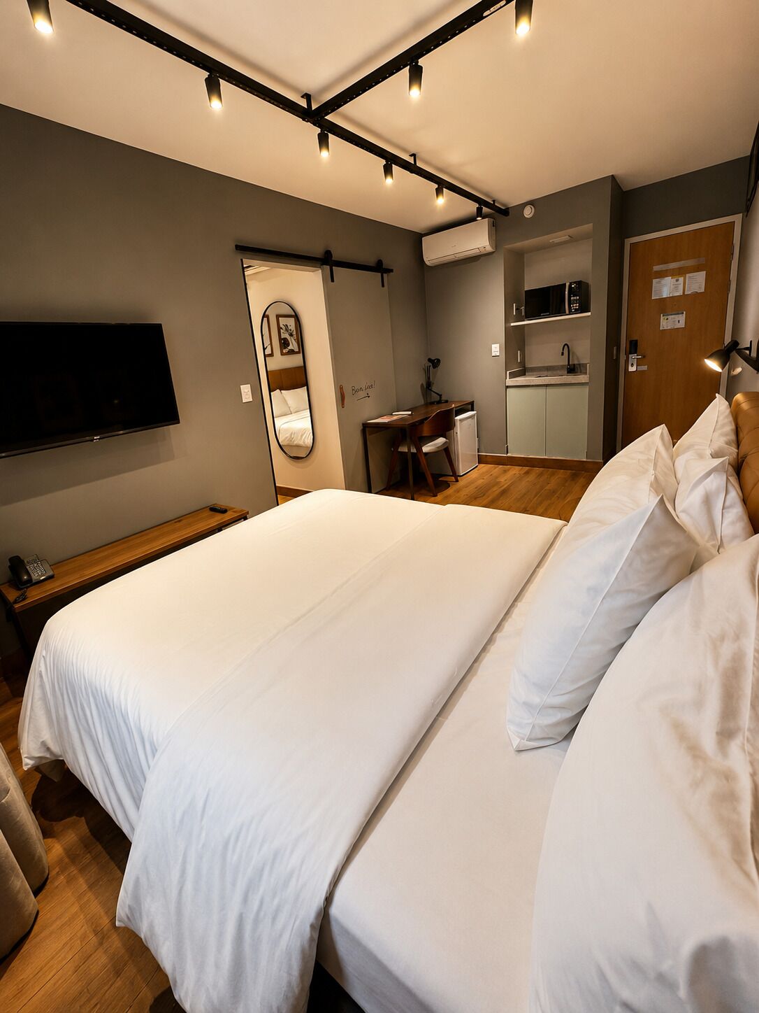 A hotel room with a large bed, a desk with a chair, a flat-screen TV, a mirror, and a kitchenette.