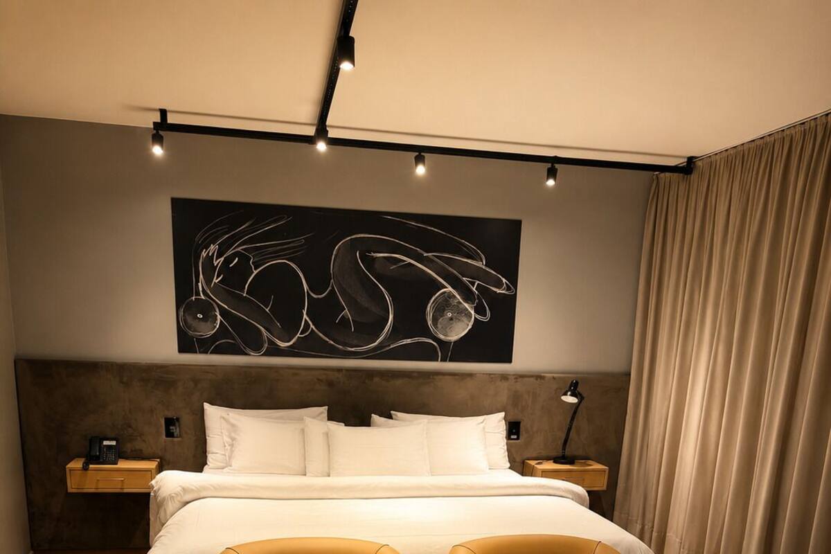 A modern hotel room with a large bed, two armchairs, a small table, a nightstand, a lamp, and a wall art piece.