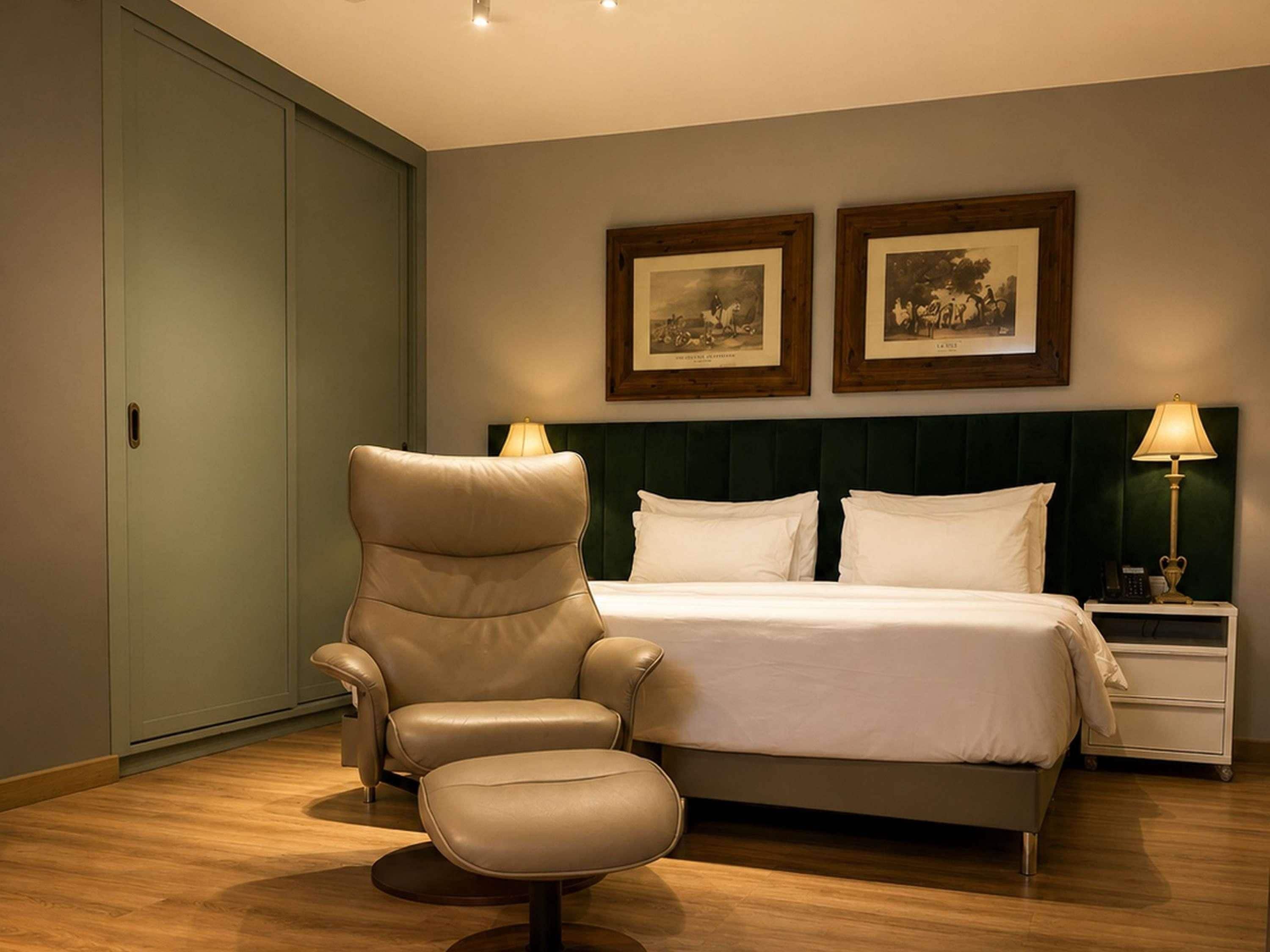 A hotel room with a bed, a chair, a nightstand, and two framed pictures on the wall.
