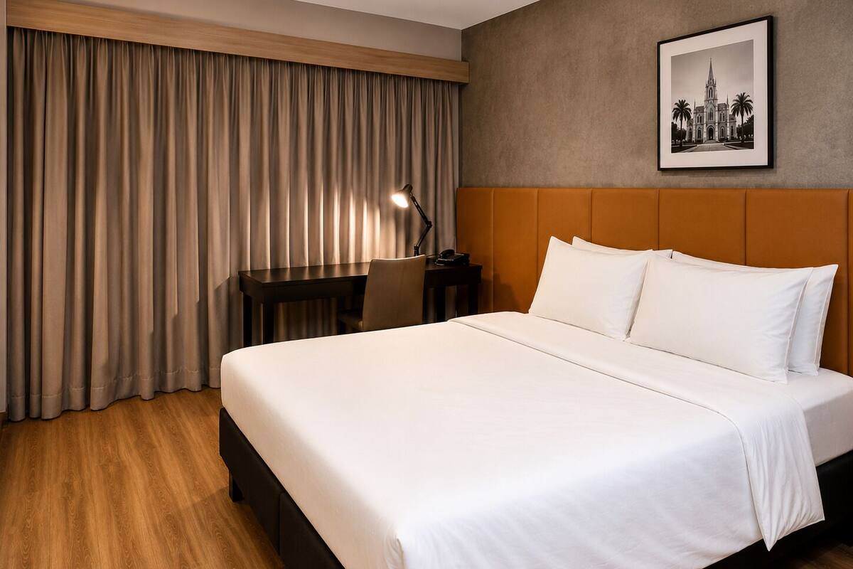 A hotel room with a bed, a desk, a chair, a lamp, and a framed picture on the wall.