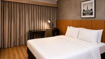 A hotel room with a bed, a desk, a chair, a lamp, and a framed picture on the wall.
