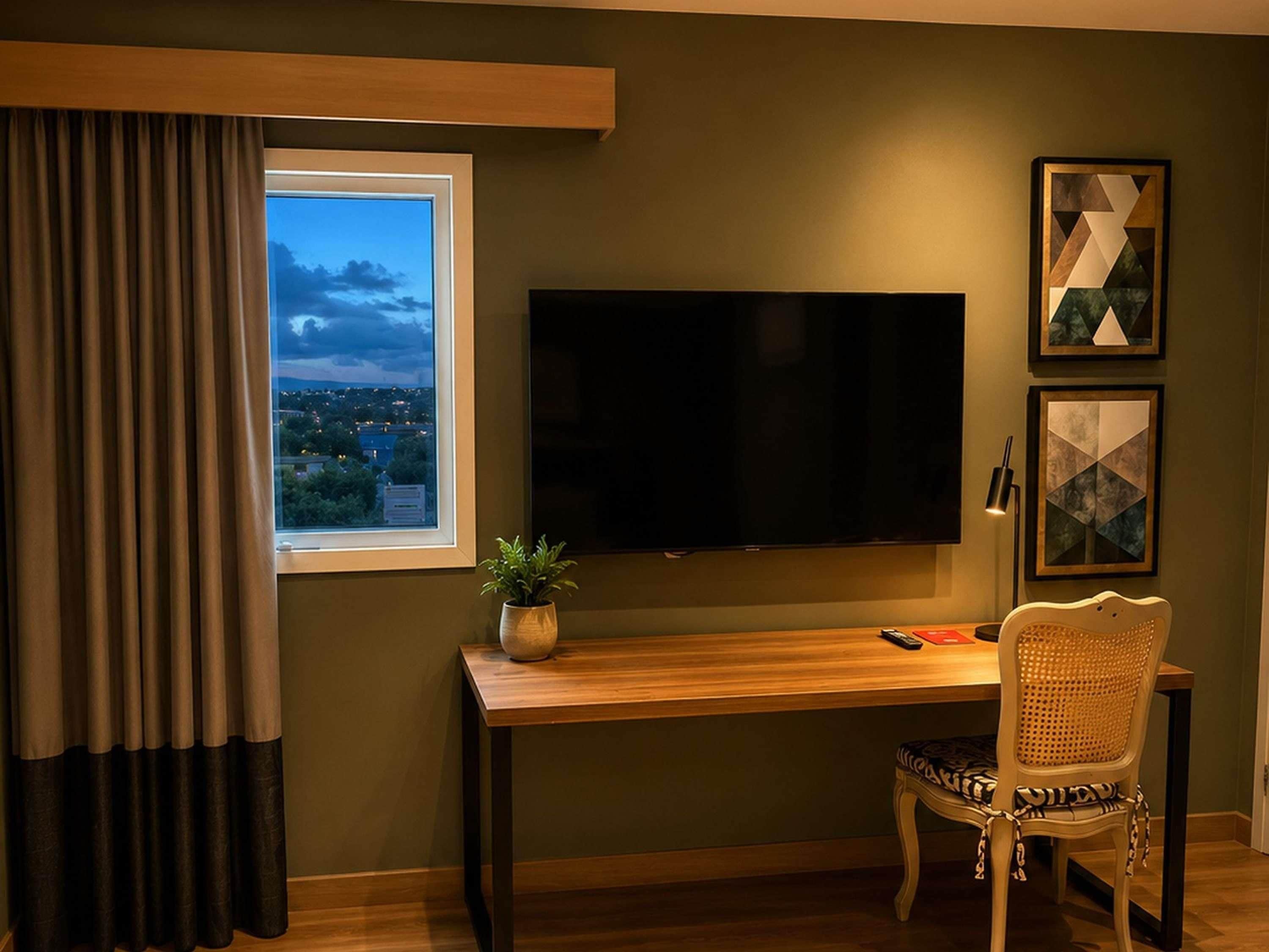 A room with a large flat-screen TV mounted on the wall, a wooden desk, a wicker chair, and a window with curtains.