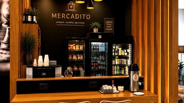 A bar area with a wooden counter, two bar stools, a refrigerator stocked with beverages, and a wall with the text 'MERCADITO' and a slogan. - ibis Catanduva (Catanduva)
