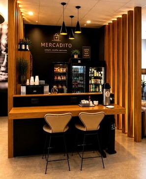 A bar area with a wooden counter, two bar stools, a refrigerator stocked with beverages, and a wall with the text 'MERCADITO' and a slogan. - ibis Catanduva (Catanduva)