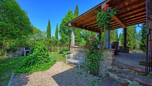 En bänk av sten under en träpergola, omgiven av klätterrosor, grönska och en klarblå himmel.