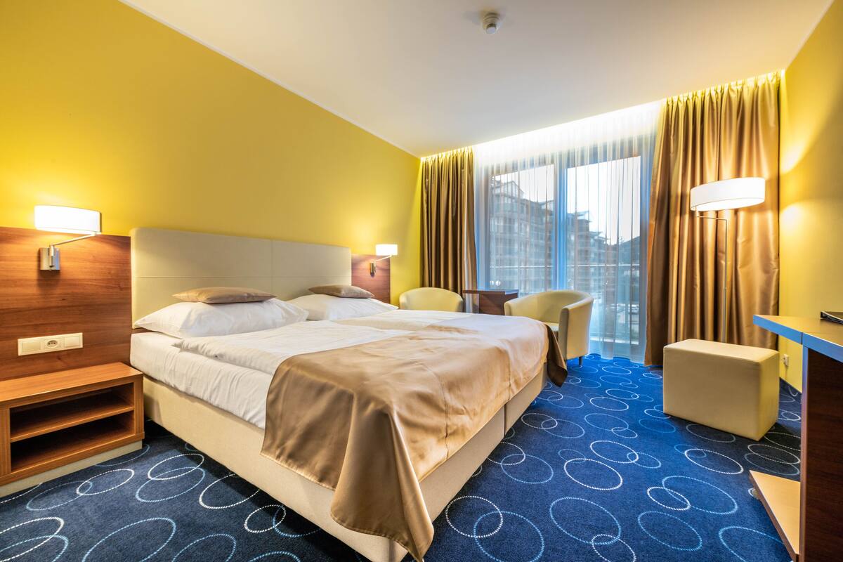 Quarto de hotel com uma cama grande, uma escrivaninha, uma cadeira e vista para a cidade pela janela.